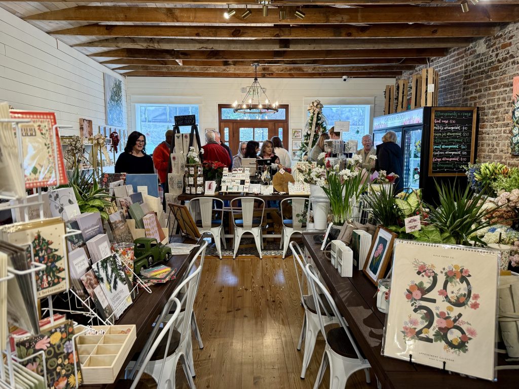 A cozy shop with wooden floors, white chairs, and tables displaying stationery and gifts; several people are browsing and conversing near the back.