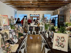 A cozy shop with wooden floors, white chairs, and tables displaying stationery and gifts; several people are browsing and conversing near the back.