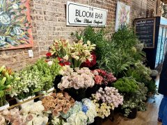 A variety of fresh flowers in buckets are displayed against a brick wall inside a flower shop with a sign reading “The Bloom Bar.”.