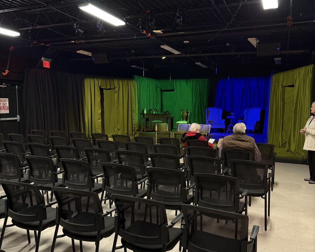 A small theater with rows of empty chairs faces a stage set with green and blue lighting. Three people are seated and one person stands at the right edge of the image.