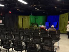A small theater with rows of empty chairs faces a stage set with green and blue lighting. Three people are seated and one person stands at the right edge of the image.