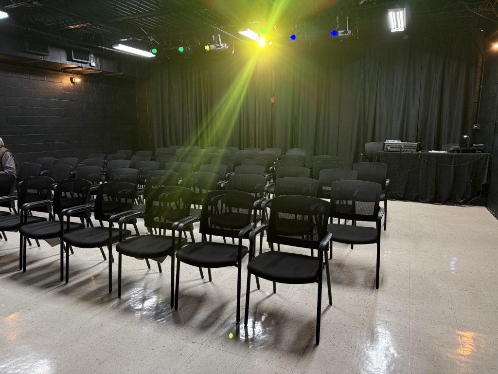 Rows of empty black chairs face a black curtain stage in a small, well-lit theater with colorful stage lights overhead.