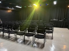 Rows of empty black chairs face a black curtain stage in a small, well-lit theater with colorful stage lights overhead.