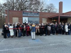A large group of people gather outside the Rena Henton Theatre and Robert’s School Community & Education Center as a ribbon is cut at an outdoor ceremony.