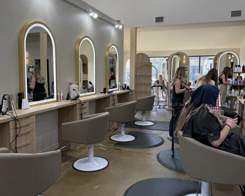 A modern hair salon with beige chairs and large mirrors. Several clients are getting their hair done by stylists. Shelves with hair products are visible in the background.