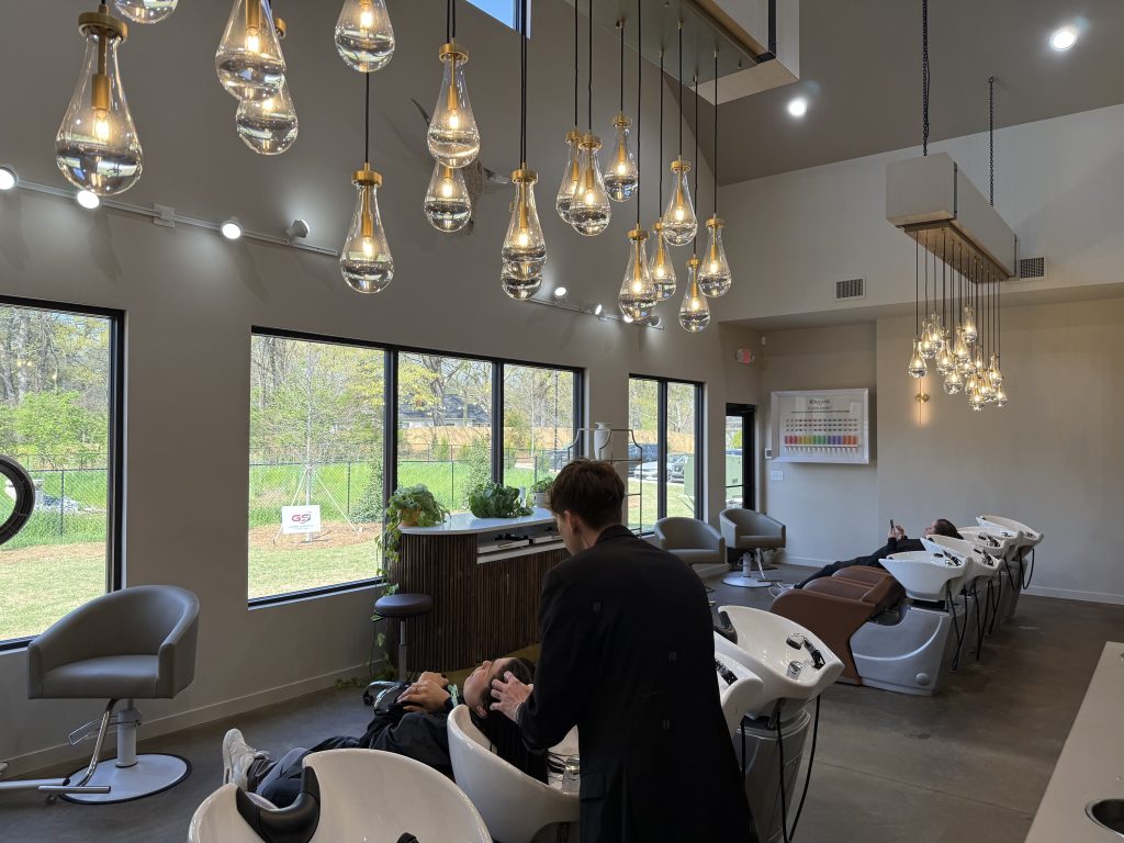 A hair salon with large windows, modern hanging lights, and a stylist washing a client's hair at a row of wash stations.