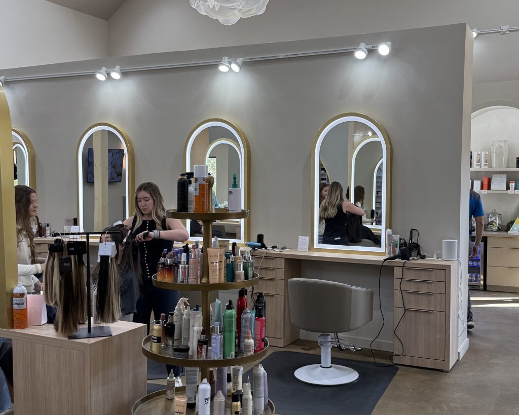 A hair salon with light wood furniture, mirrors, and shelves stocked with hair products. A stylist is working with a client on the left, while another woman sits in a chair facing a mirror.