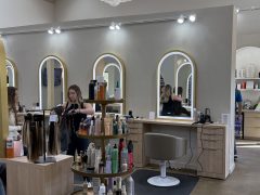 A hair salon with light wood furniture, mirrors, and shelves stocked with hair products. A stylist is working with a client on the left, while another woman sits in a chair facing a mirror.