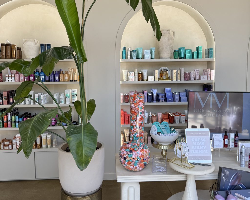 A modern beauty boutique interior with shelves of skincare products, a large potted plant, a glass jar filled with colorful capsules, and a sign that reads “GUESS HOW MANY FUSIO’S.”.