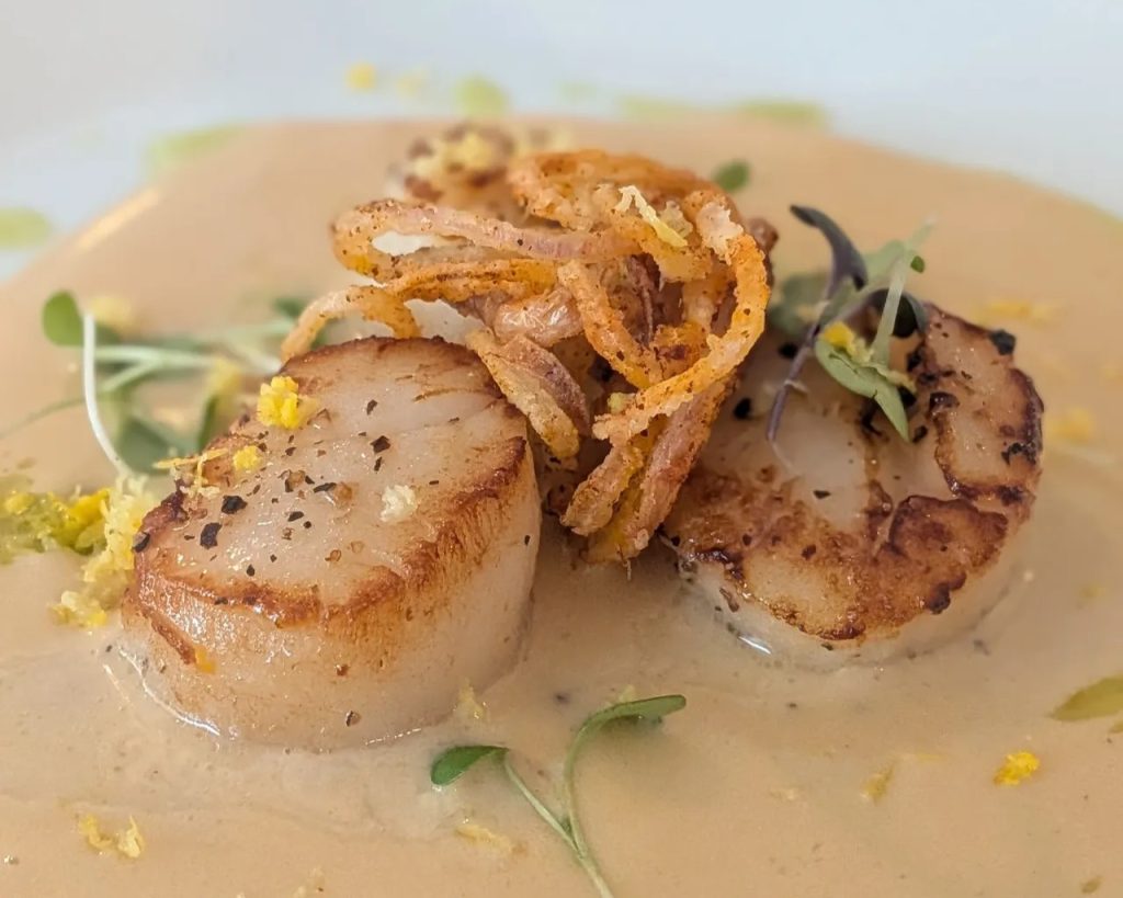 Two seared scallops with cracked pepper on a creamy sauce, topped with crispy fried onions and microgreens.