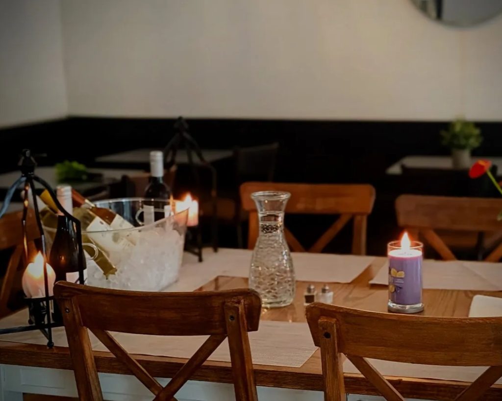 A wooden dining table is set with placemats, a carafe, candles, and a bucket of wine bottles on ice. Four chairs surround the table in a softly lit restaurant.