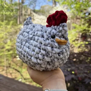 A hand holds a crocheted chicken with a gray and white body, black eyes, a red comb, and an orange beak. Green trees and blue sky are visible in the background.