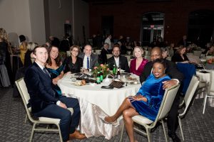 Eight people in formal attire sit around a round table at an indoor event, smiling for a group photo. The table has drinks, food, and floral centerpieces. Other guests are visible in the background.