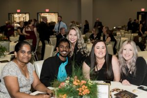 Four people sit at a round table with floral centerpiece, smiling at the camera. Other attendees are seated and standing in the background at an indoor event.