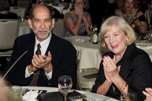 An older man in a suit and a woman in black clothing are seated at a round table, clapping at an event with other people in the background.