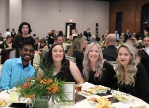 A group of five people sit at a round table with food and floral centerpieces at a formal event in a large banquet hall.