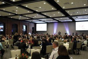 A large group of people seated at round tables during a formal event in a spacious banquet hall, with a speaker at a podium and presentation screens visible.