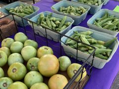 Green tomatoes, cartons of okra, and jars of honey are displayed on a purple tablecloth at an outdoor market stand.