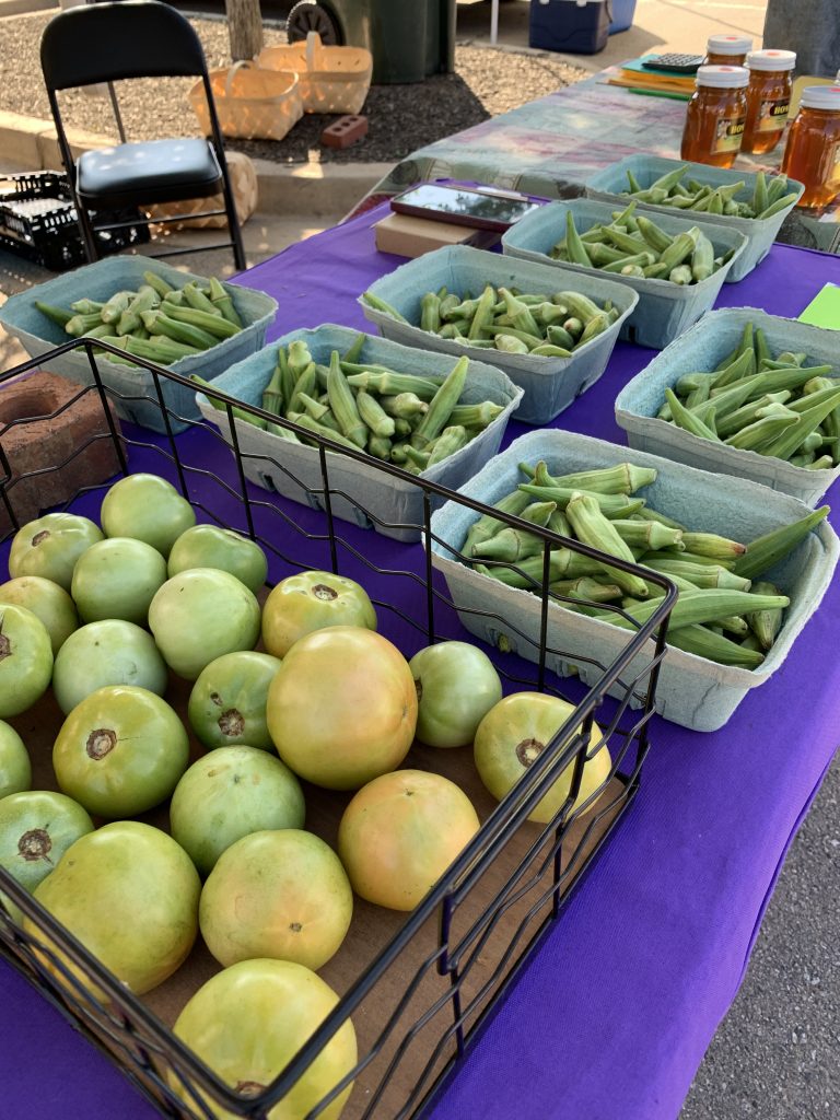 Green tomatoes, cartons of okra, and jars of honey are displayed on a purple tablecloth at an outdoor market stand.