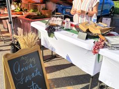 A woman stands behind a farmers market booth displaying bread, eggs, and pasta. A chalkboard sign in front lists "Amy's Lavata Pasta." A parked car is visible in the background.