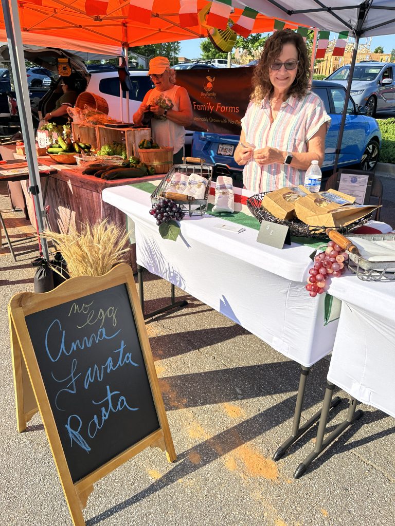 A woman stands behind a farmers market booth displaying bread, eggs, and pasta. A chalkboard sign in front lists 
