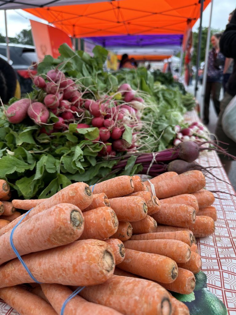 Bundles of carrots, radishes, and beets displayed on a table at an outdoor farmers market under orange and purple canopy tents.