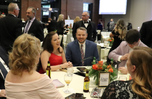 People dressed in formal attire sit around a table with drinks and flowers at an indoor event, engaging in conversation and smiling.