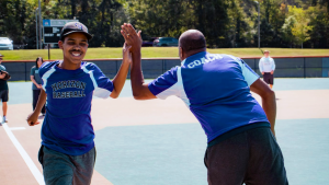 A young baseball player and a coach, both in blue uniforms, high-five on a baseball field while others watch in the background.