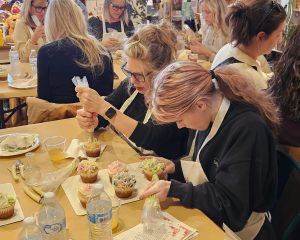 A group of people at a table decorate cupcakes with icing using piping bags during a workshop or class.