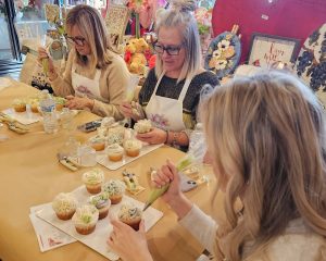Three women sit at a table decorating cupcakes with icing bags. Art supplies, decorated cupcakes, water bottles, and floral crafts are visible on the table.