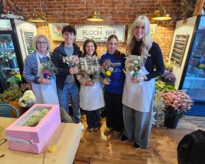 Five people wearing aprons stand in a flower shop holding floral arrangements, smiling at the camera. A pink box and flower displays are visible in the background.