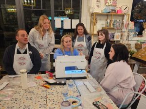 A group of people wearing aprons watch a woman demonstrate sewing on a white sewing machine in a craft room.