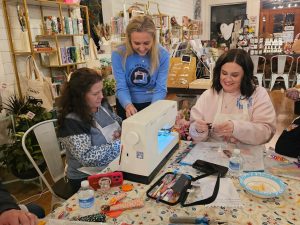 Three women work together at a table with a sewing machine, crafting supplies, and snacks in a cozy, well-lit room.