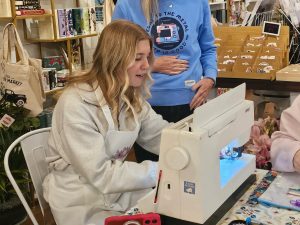 A woman wearing a white apron uses a sewing machine at a table while another person stands nearby with a hand on their stomach.