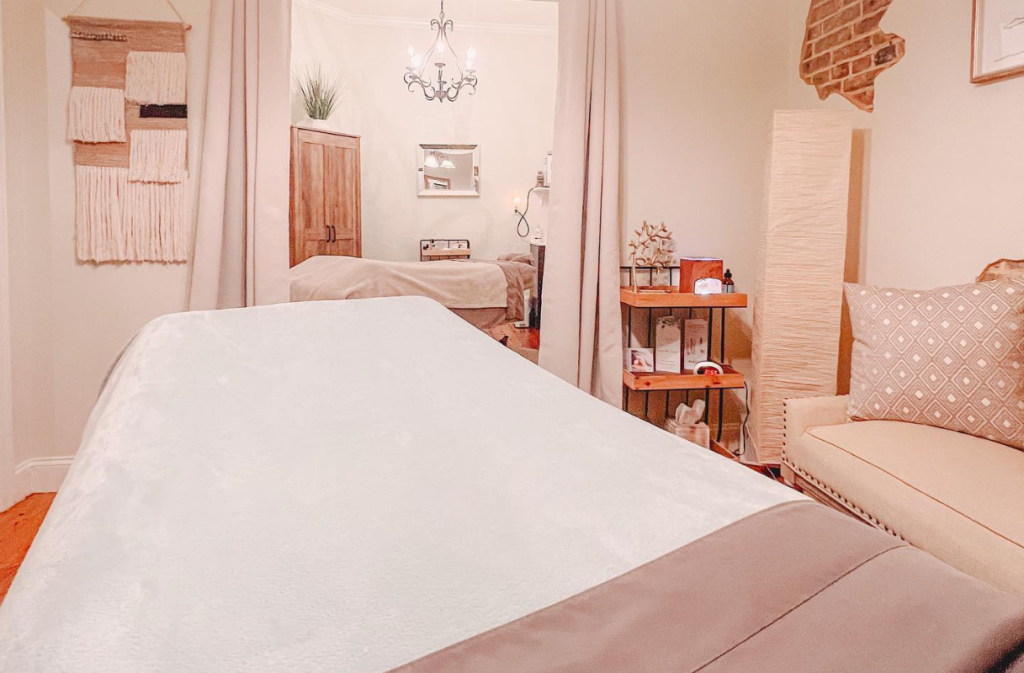 A neatly made massage table in a softly lit room with beige walls, decorative accents, and a small seating area.