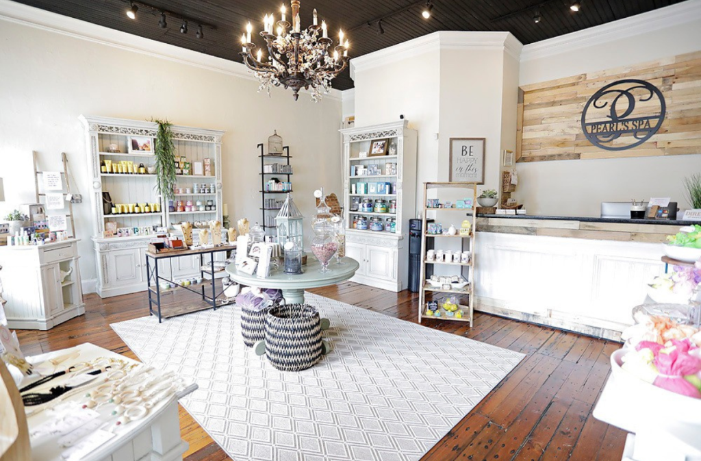 A boutique shop interior with shelves of products, a round display table, a chandelier, wooden floors, and a counter with a sign reading “P.”.