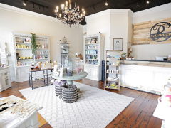 A boutique shop interior with shelves of products, a round display table, a chandelier, wooden floors, and a counter with a sign reading “P.”.