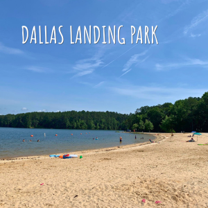 Welcome to Acworth, Georgia—a sandy beach at Dallas Landing Park with people swimming and playing near the water; trees line the background under a clear blue sky.