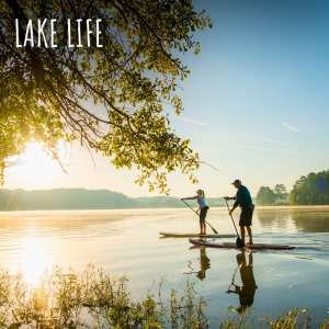 Two people stand-up paddleboarding on a calm lake at sunrise, with overhanging tree branches and the words "Lake Life" in the top left corner. You're Welcome in Acworth to experience serene mornings like this.