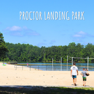 Two people walk on a sandy beach near a volleyball court and a lake, surrounded by trees under a clear blue sky. Text reads "Proctor Landing Park. Welcome to Acworth Georgia.