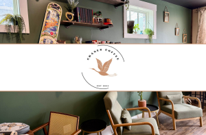 A cozy coffee shop interior with green walls, wooden chairs, small tables, plants, and wall shelves; the Cranes Coffee logo is centered in the middle of the image.