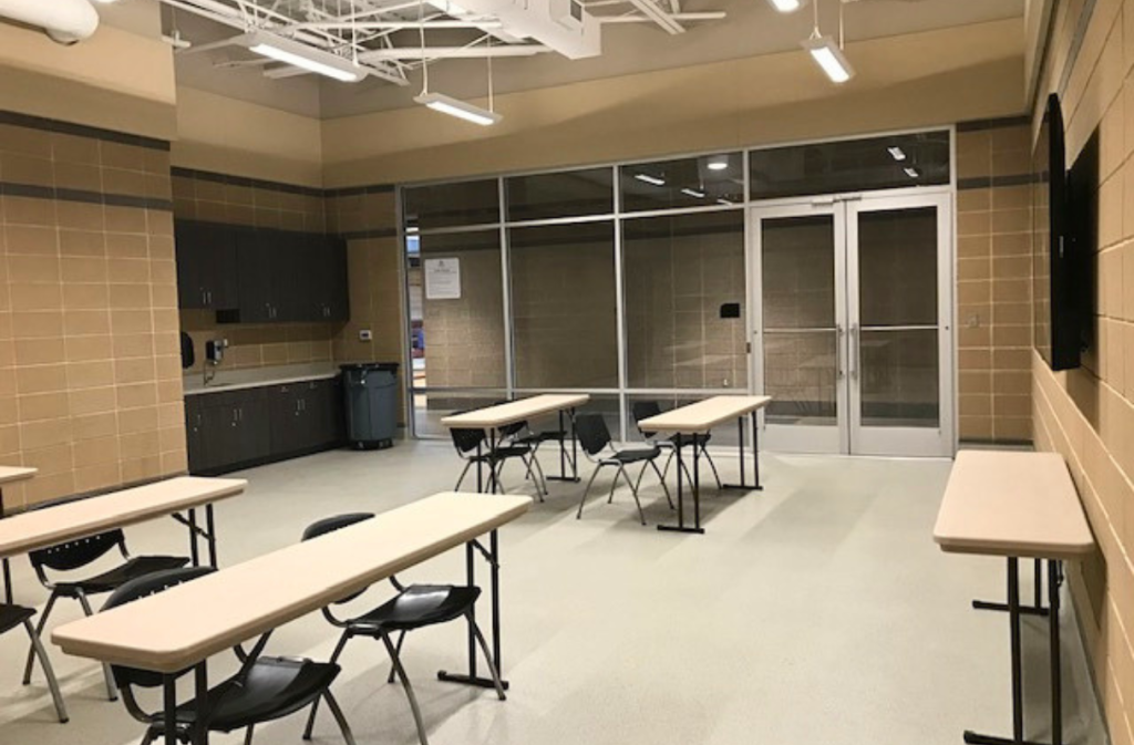 A classroom with beige walls, several rectangular tables and black chairs, cabinets along the back wall, and glass double doors at the front.