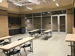 A classroom with beige walls, several rectangular tables and black chairs, cabinets along the back wall, and glass double doors at the front.