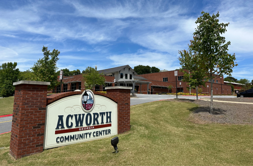 Acworth Community Center building with a sign in the foreground on a grassy area, under a blue sky with scattered clouds.