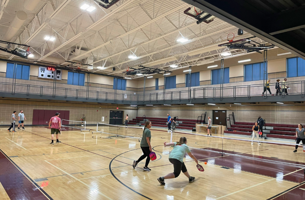 People play pickleball on several courts inside a gymnasium, with others watching from bleachers and a balcony. The gym has high ceilings and bright lighting.