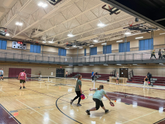 People play pickleball on several courts inside a gymnasium, with others watching from bleachers and a balcony. The gym has high ceilings and bright lighting.