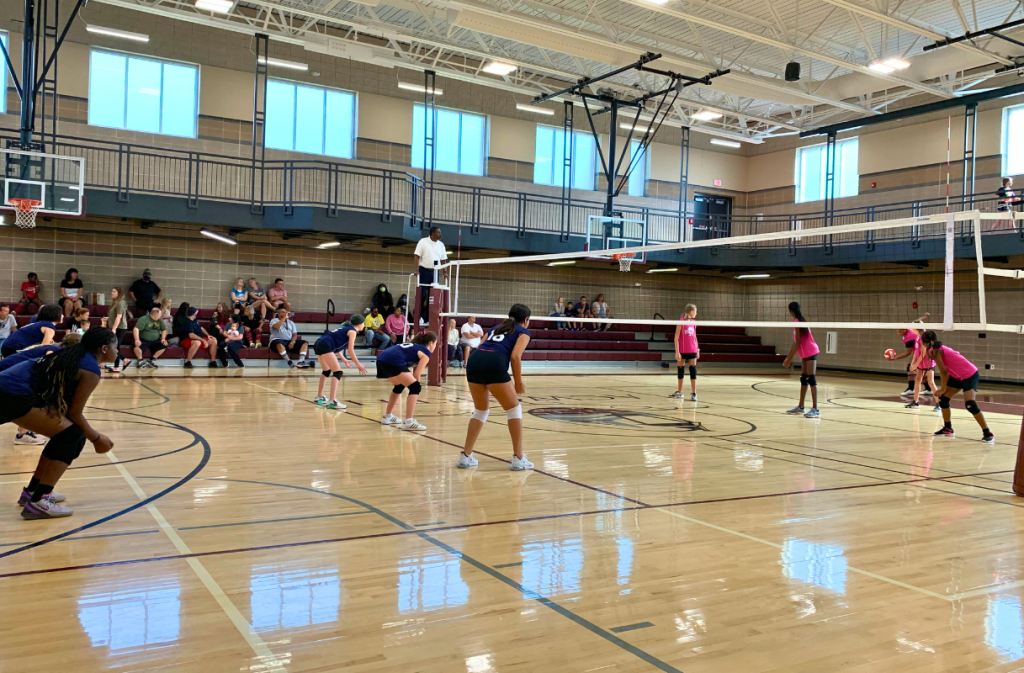 Two girls' volleyball teams face each other in a gymnasium, with one team preparing to serve and spectators watching from bleachers.