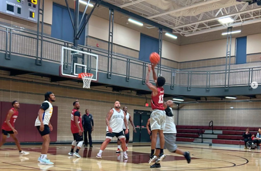 A basketball player in a red jersey jumps to take a shot while other players in white and red jerseys watch on an indoor court.