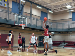 A basketball player in a red jersey jumps to take a shot while other players in white and red jerseys watch on an indoor court.