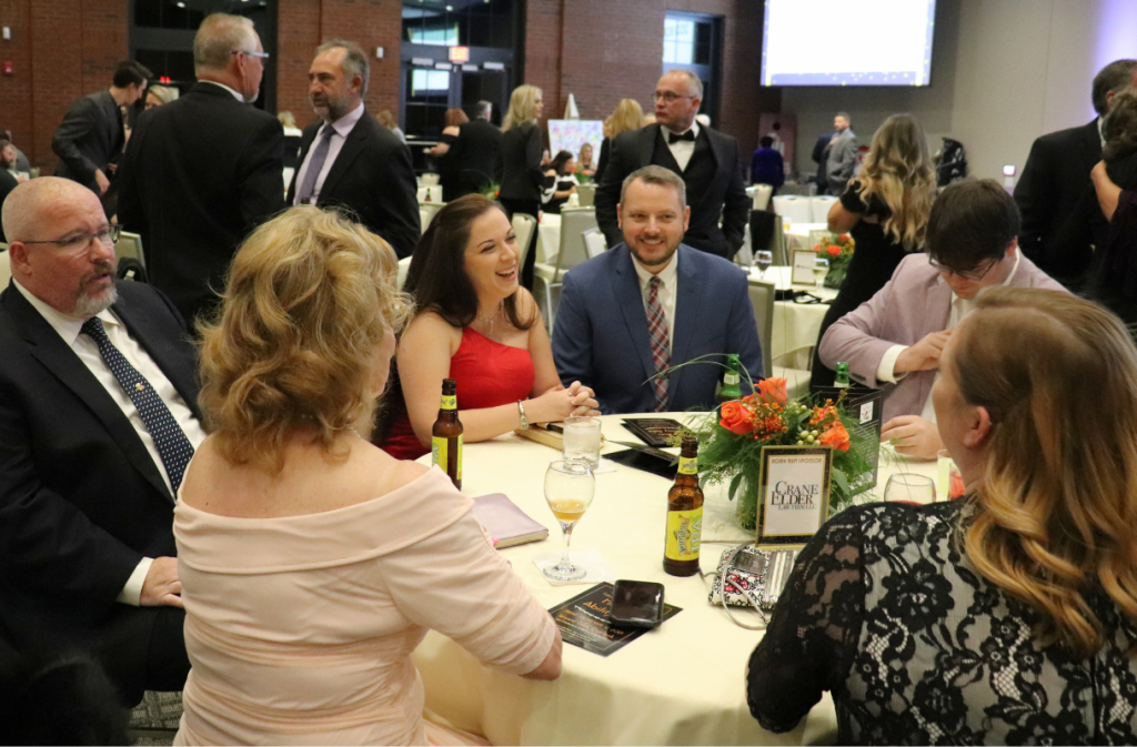 People in formal attire sit at a round table with drinks and programs at a banquet or gala event, with more guests and tables visible in the background.
